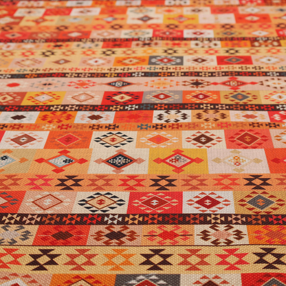 A geometric woven fabric featuring horizontal rows of Aztec and tribal-inspired patterns in warm earthy tones of orange, red, yellow, beige, brown, and dark charcoal.