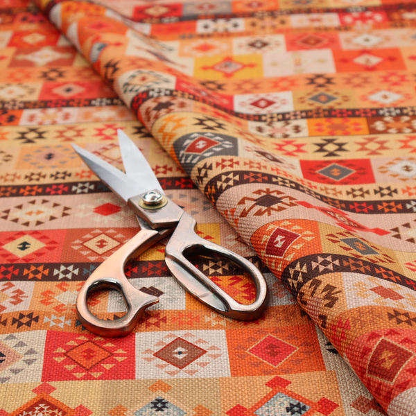 A geometric woven fabric featuring horizontal rows of Aztec and tribal-inspired patterns in warm earthy tones of orange, red, yellow, beige, brown, and dark charcoal.