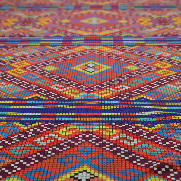 A vibrant geometric fabric featuring a detailed pixelated pattern of large interlocking diamonds in red, blue, purple, yellow, and turquoise, separated by horizontal striped bands of multi-coloured dots on a dark blue background, reminiscent of traditional beadwork or woven textile art.