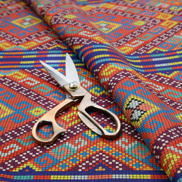 A vibrant geometric fabric featuring a detailed pixelated pattern of large interlocking diamonds in red, blue, purple, yellow, and turquoise, separated by horizontal striped bands of multi-coloured dots on a dark blue background, reminiscent of traditional beadwork or woven textile art.