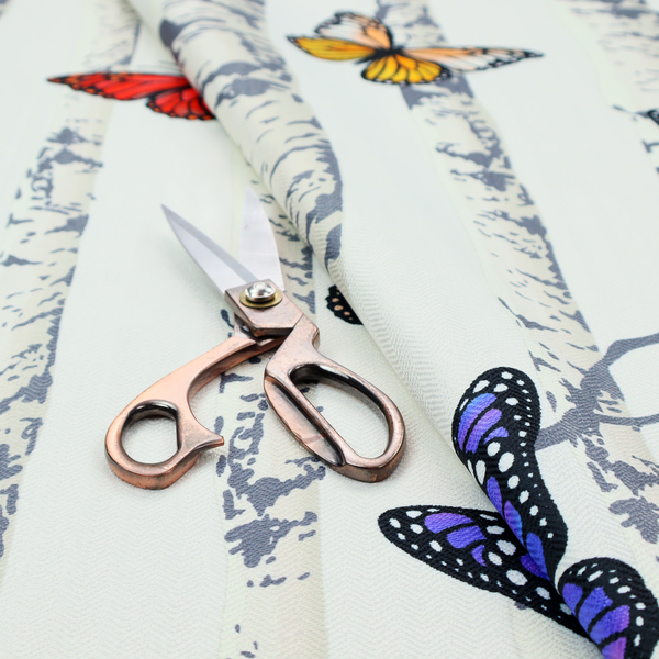A light-coloured fabric featuring a pattern of vertical grey and white birch tree trunks interspersed with colourful butterflies in shades of red, orange, yellow, blue, and purple.