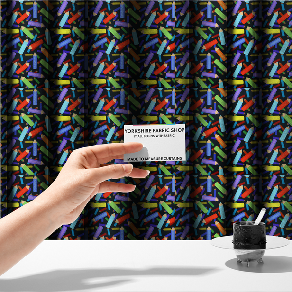 The fabric used to create these made-to-measure curtains is a black fabric featuring a playful, repeating pattern of coloured pencils in various shades including red, yellow, light blue, green, purple, and orange scattered diagonally across the background.