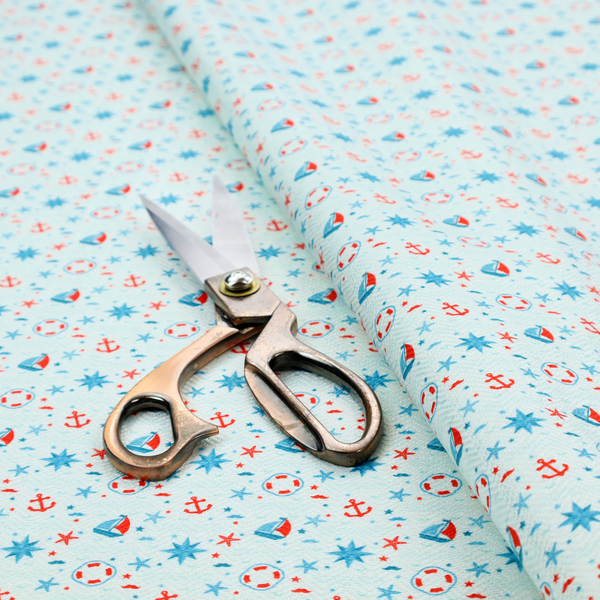 Light blue fabric featuring a small-scale repeating nautical pattern of red anchors, lifebuoys, and sailboats, interspersed with blue and red stars.