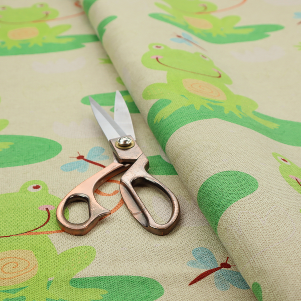 A repeating pattern fabric featuring cute cartoon green frogs sitting on bright green lily pads, interspersed with blue and red dragonflies and white water lilies on a light cream background.