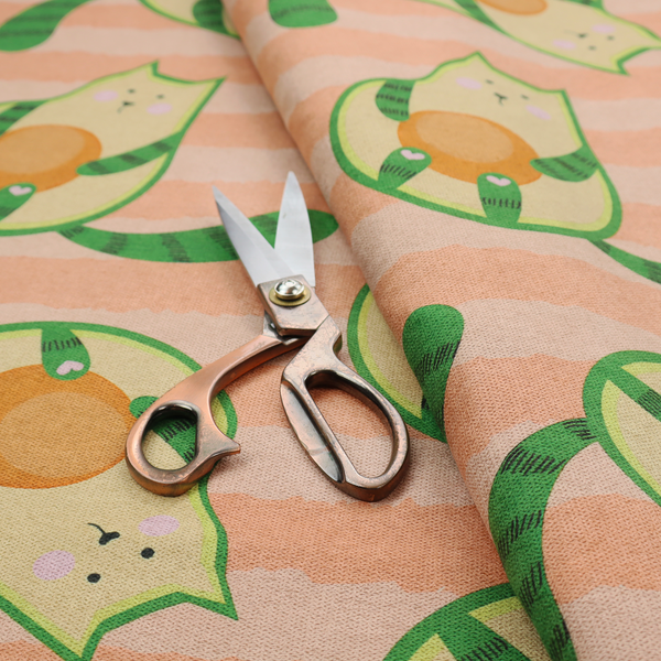 A playful repeating pattern fabric featuring whimsical illustrated "avocatos"—cats shaped like avocado halves with green striped tails and orange pits—over a light peach background with horizontal orange stripes.