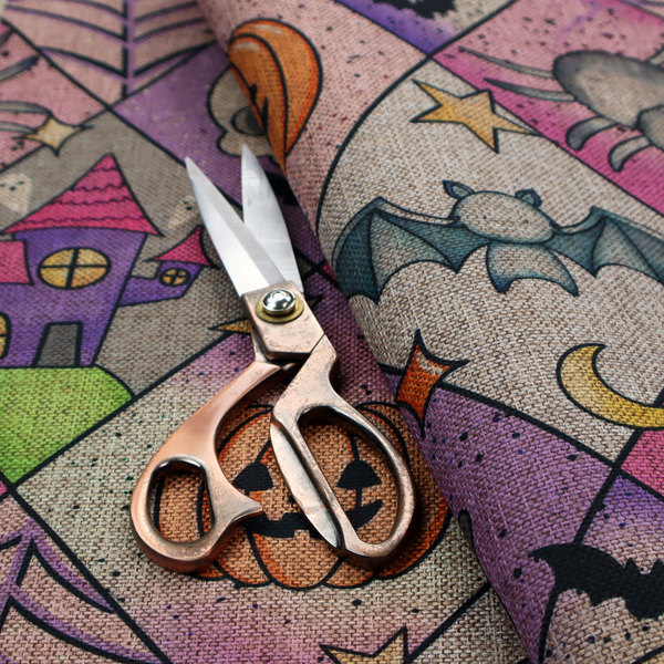 A Halloween-themed harlequin pattern fabric with diamond-shaped sections in purple, pink, and beige, featuring cartoon illustrations of Jack-o'-lanterns, a witch's cat, ghosts, bats, skulls, spiders, stars, and crescent moons.