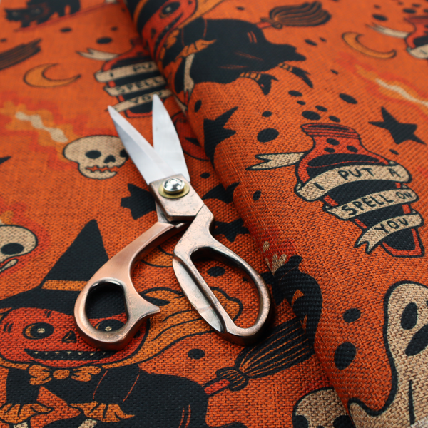 An orange Halloween fabric patterned with various black illustrations, including a witch with a pumpkin head flying on a broomstick, a black cat, ghosts, skulls, potion bottles with the text "I put a spell on you," bats, cauldrons, and stars.