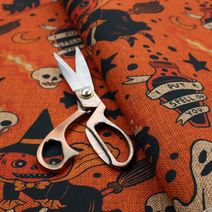 An orange Halloween fabric patterned with various black illustrations, including a witch with a pumpkin head flying on a broomstick, a black cat, ghosts, skulls, potion bottles with the text "I put a spell on you," bats, cauldrons, and stars.