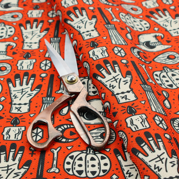 An orange Halloween fabric patterned with repeating black and white illustrations of patterned pumpkins, mystical hands with symbols, witch hats with crescent moons, brooms, candles, bones, and planchettes.