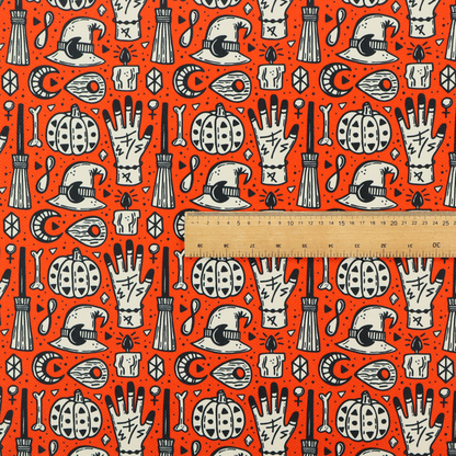 An orange Halloween fabric patterned with repeating black and white illustrations of patterned pumpkins, mystical hands with symbols, witch hats with crescent moons, brooms, candles, bones, and planchettes.