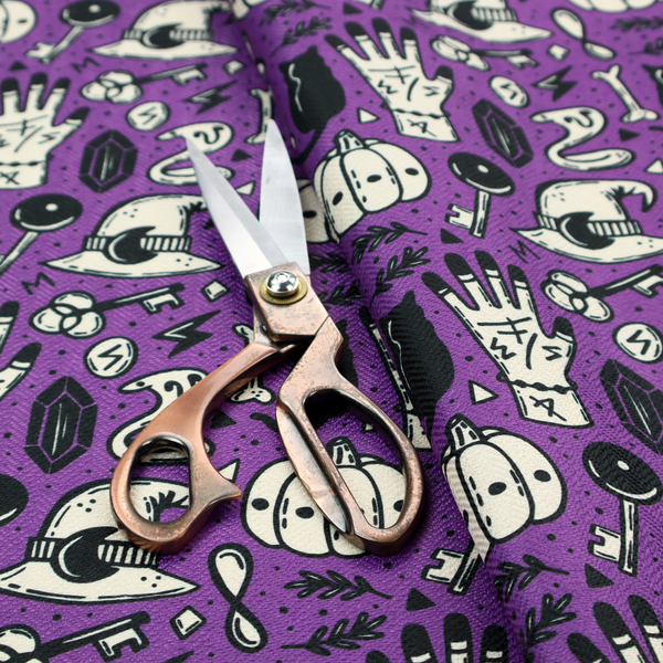 A purple Halloween fabric patterned with repeating black and cream illustrations of pumpkins, witch hats, black cats, mystical marked hands, snakes, keys, bones, and crystals.