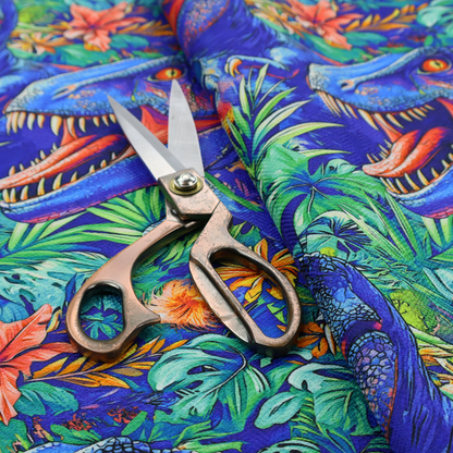 A vibrant, repeating pattern on a rich blue background featuring blue-and-purple Tyrannosaurus rex heads with open jaws and sharp teeth, surrounded by lush green tropical foliage, orange flowers, and plants.