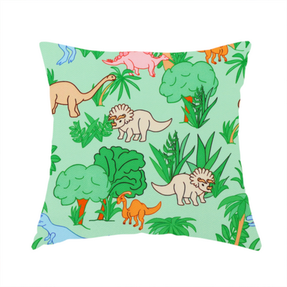 The fabric used to create this handmade cushion is a repeating pattern on a light green background featuring colourful illustrated dinosaurs—including blue T-Rexes, pink Stegosauruses, beige Triceratopses, and orange sauropods—surrounded by green palm trees, bushes, and tropical foliage.