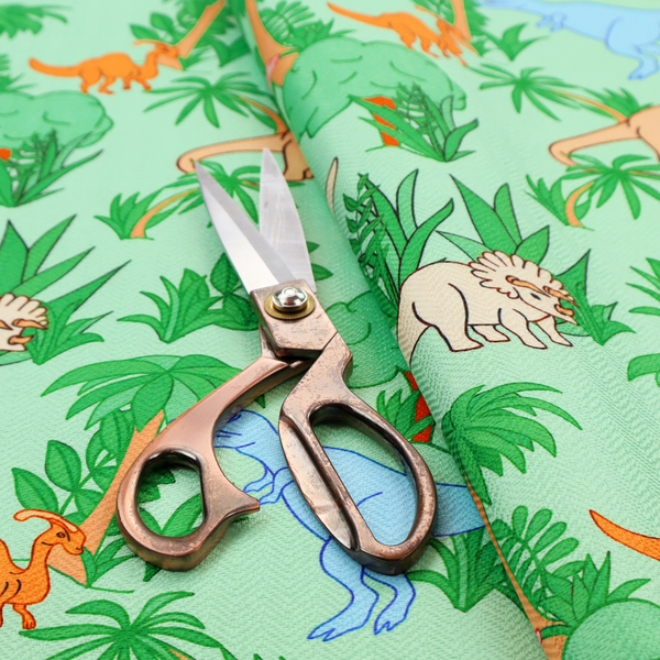 A repeating pattern on a light green background featuring colourful illustrated dinosaurs—including blue T-Rexes, pink Stegosauruses, beige Triceratopses, and orange sauropods—surrounded by green palm trees, bushes, and tropical foliage.