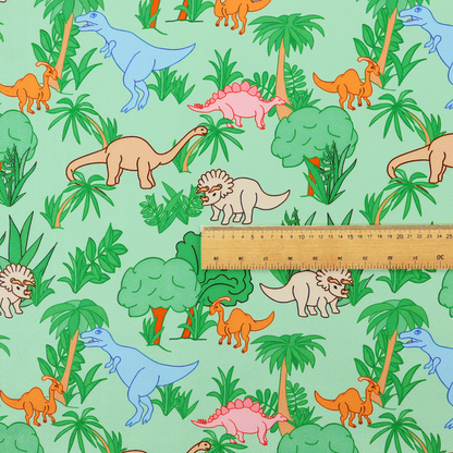 A repeating pattern on a light green background featuring colourful illustrated dinosaurs—including blue T-Rexes, pink Stegosauruses, beige Triceratopses, and orange sauropods—surrounded by green palm trees, bushes, and tropical foliage.