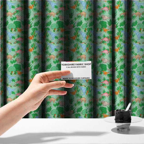 The fabric used to create these made-to-measure curtains is a repeating pattern on a light green background featuring colourful illustrated dinosaurs—including blue T-Rexes, pink Stegosauruses, beige Triceratopses, and orange sauropods—surrounded by green palm trees, bushes, and tropical foliage.