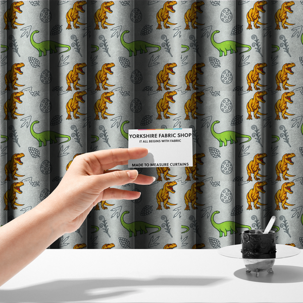 The fabric used to create these made-to-measure curtains is a repeating pattern on a light grey-blue background featuring orange Tyrannosaurus rex dinosaurs, a bright green long-necked sauropod, speckled dinosaur eggs, pinecones, footprints, and leafy sprigs.