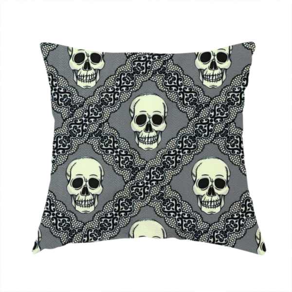 The fabric used to create this handmade cushion is a repeating pattern on a grey background featuring off-white human skulls arranged in a grid, separated by diagonal intersecting bands of ornate black lace with intricate scrollwork.