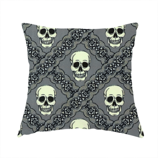 The fabric used to create this handmade cushion is a repeating pattern on a grey background featuring off-white human skulls arranged in a grid, separated by diagonal intersecting bands of ornate black lace with intricate scrollwork.