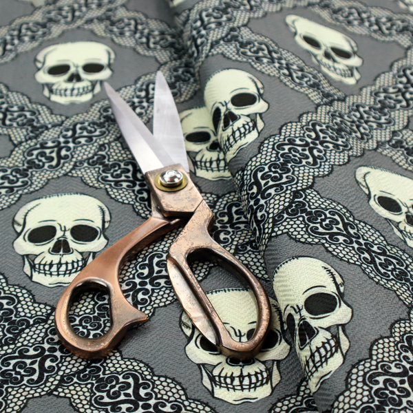 A repeating pattern on a grey background featuring off-white human skulls arranged in a grid, separated by diagonal intersecting bands of ornate black lace with intricate scrollwork.