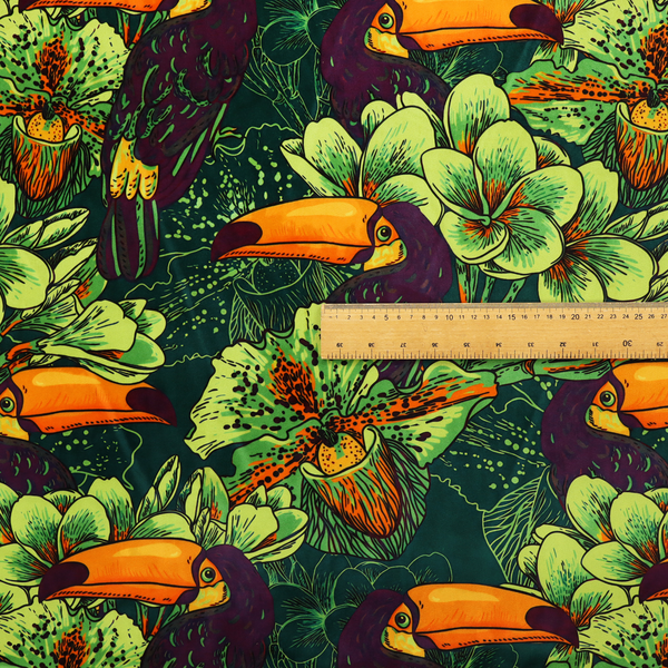 A dark green fabric densely patterned with toucans featuring large orange-and-yellow beaks, surrounded by glowing light green and orange tropical flowers.   