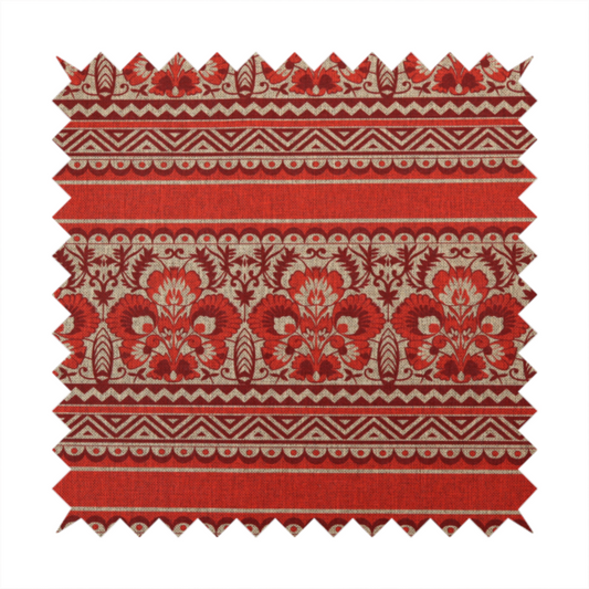 A fabric featuring a folkloric red and beige pattern with horizontal bands of floral motifs, chevrons, and dotted borders.