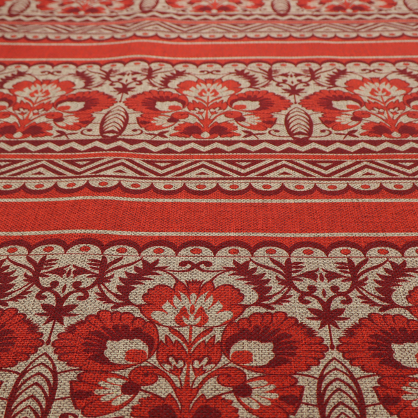 A fabric featuring a folkloric red and beige pattern with horizontal bands of floral motifs, chevrons, and dotted borders.