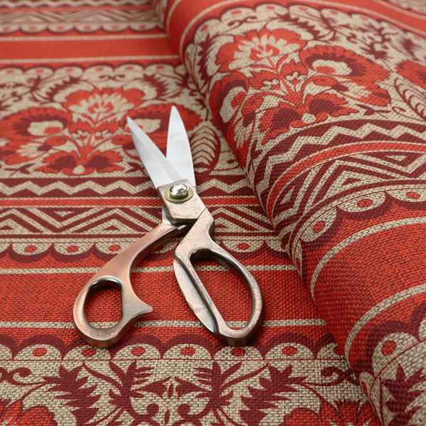 A fabric featuring a folkloric red and beige pattern with horizontal bands of floral motifs, chevrons, and dotted borders.