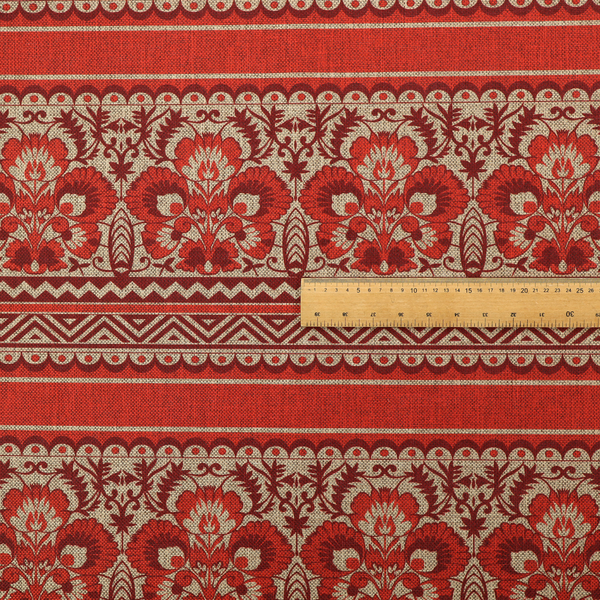 A fabric featuring a folkloric red and beige pattern with horizontal bands of floral motifs, chevrons, and dotted borders.