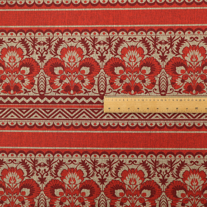 A fabric featuring a folkloric red and beige pattern with horizontal bands of floral motifs, chevrons, and dotted borders.