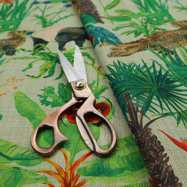 A green and beige fabric patterned with hand-drawn jungle illustrations of alligators, a tapir, tropical trees, and vibrant flowers featuring an organic style.