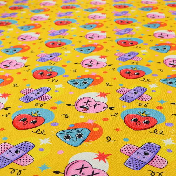 Yellow fabric featuring a playful, quirky pattern with cartoon-style graphics including smiling hearts with crowns, cute diamonds, cartoon bandages, and wacky faces with X eyes on a bright yellow background.   
