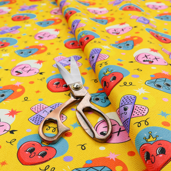 Yellow fabric featuring a playful, quirky pattern with cartoon-style graphics including smiling hearts with crowns, cute diamonds, cartoon bandages, and wacky faces with X eyes on a bright yellow background.   