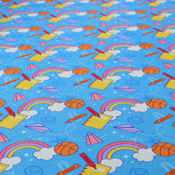 Blue fabric featuring a playful back-to-school pattern with colourful rainbows, basketballs, yellow notebooks with red pencils, purple paper airplanes, and school supplies scattered across a bright blue background.