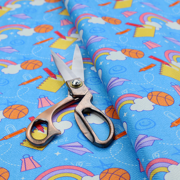 Blue fabric featuring a playful back-to-school pattern with colourful rainbows, basketballs, yellow notebooks with red pencils, purple paper airplanes, and school supplies scattered across a bright blue background.