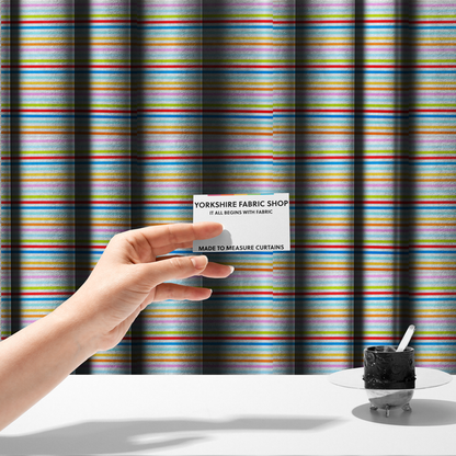 The fabric used to create these made-to-measure curtains is a white fabric patterned with horizontal, hand-drawn multi-coloured stripes in pink, blue, green, red, yellow, and orange that feature a slight, organic waviness.   