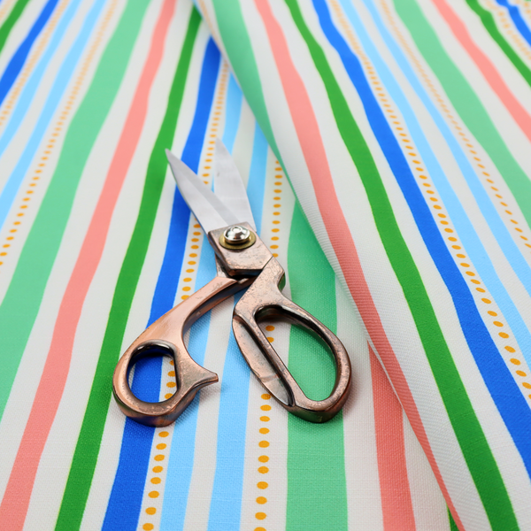 A white fabric patterned with vertical, hand-drawn multi-coloured stripes in green, blue, pink, and light blue alongside dotted lines that feature an organic waviness.