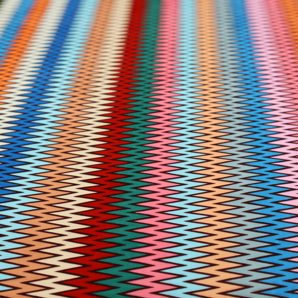A vibrant fabric pattern featuring tight vertical rows of colourful zigzag lines in shades of orange, pink, blue, red, green, and beige.