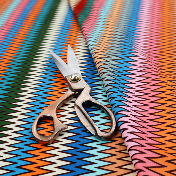 A vibrant fabric pattern featuring tight vertical rows of colourful zigzag lines in shades of orange, pink, blue, red, green, and beige.