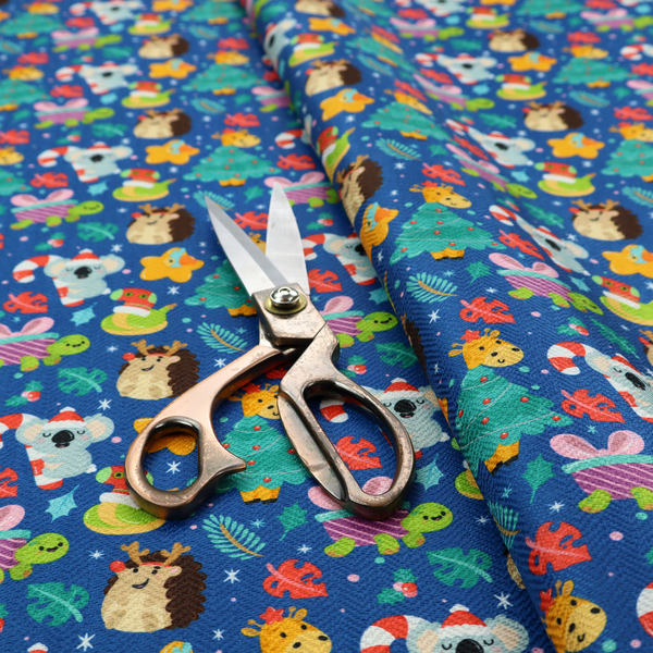 A repeating festive Christmas print on a royal blue background featuring cute cartoon holiday characters, including koalas wearing Santa hats, hedgehogs with antlers, giraffes, Christmas trees, wrapped presents, and stars.   