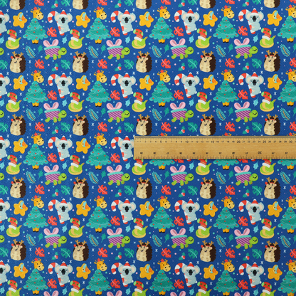 A repeating festive Christmas print on a royal blue background featuring cute cartoon holiday characters, including koalas wearing Santa hats, hedgehogs with antlers, giraffes, Christmas trees, wrapped presents, and stars.   
